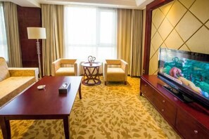 A hotel room with a sofa, two armchairs, a small round table, a TV on a wooden cabinet, and a patterned carpet.
