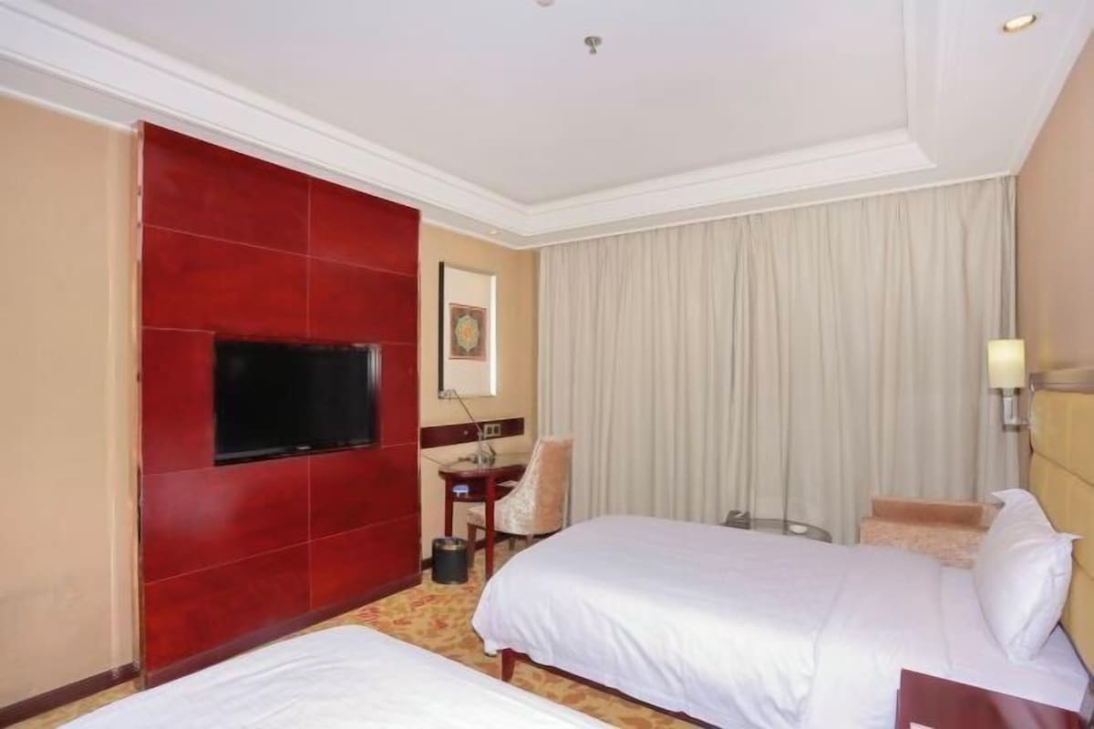 A hotel room with two beds, a TV, a desk, a chair, and a large window with curtains.