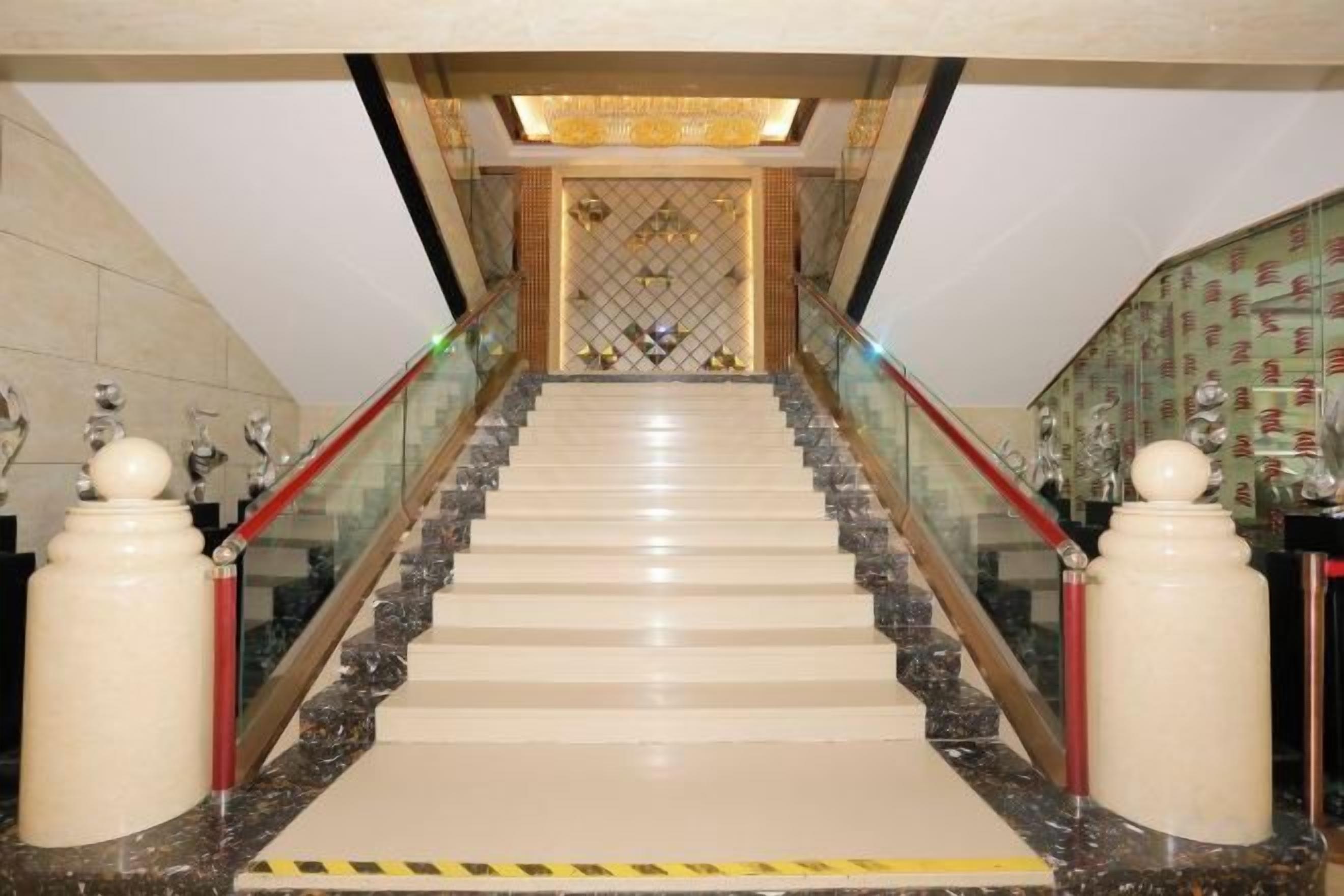 A grand staircase with marble steps and glass railings, leading to a decorative area with a patterned wall and a chandelier.