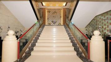 A grand staircase with marble steps and glass railings, leading to a decorative area with a patterned wall and a chandelier.