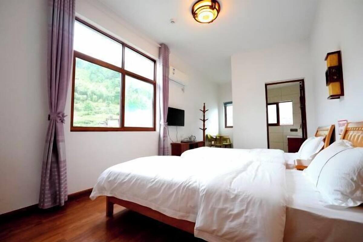 A room with a large bed, a window with curtains, a flat-screen TV, a mirror, and a wall-mounted light fixture.