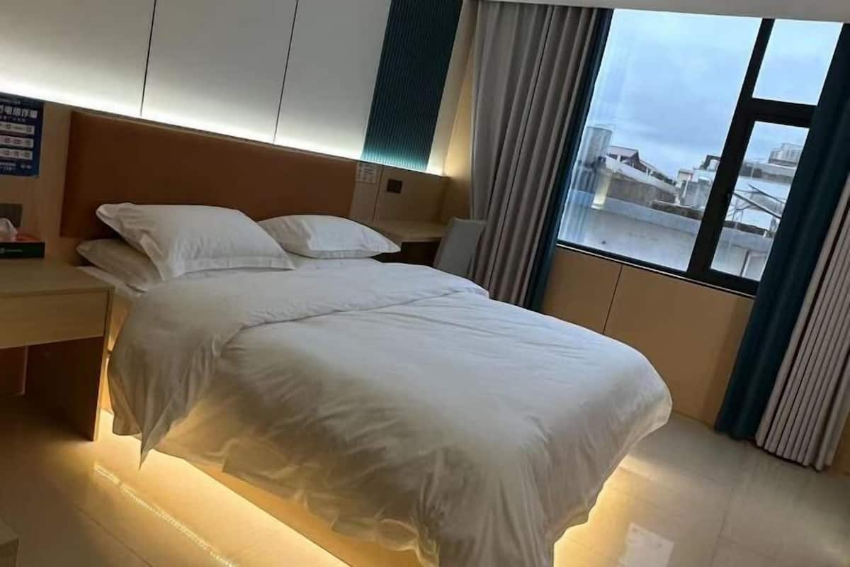 A hotel room with a bed, a nightstand, a window with curtains, and a view of buildings outside.