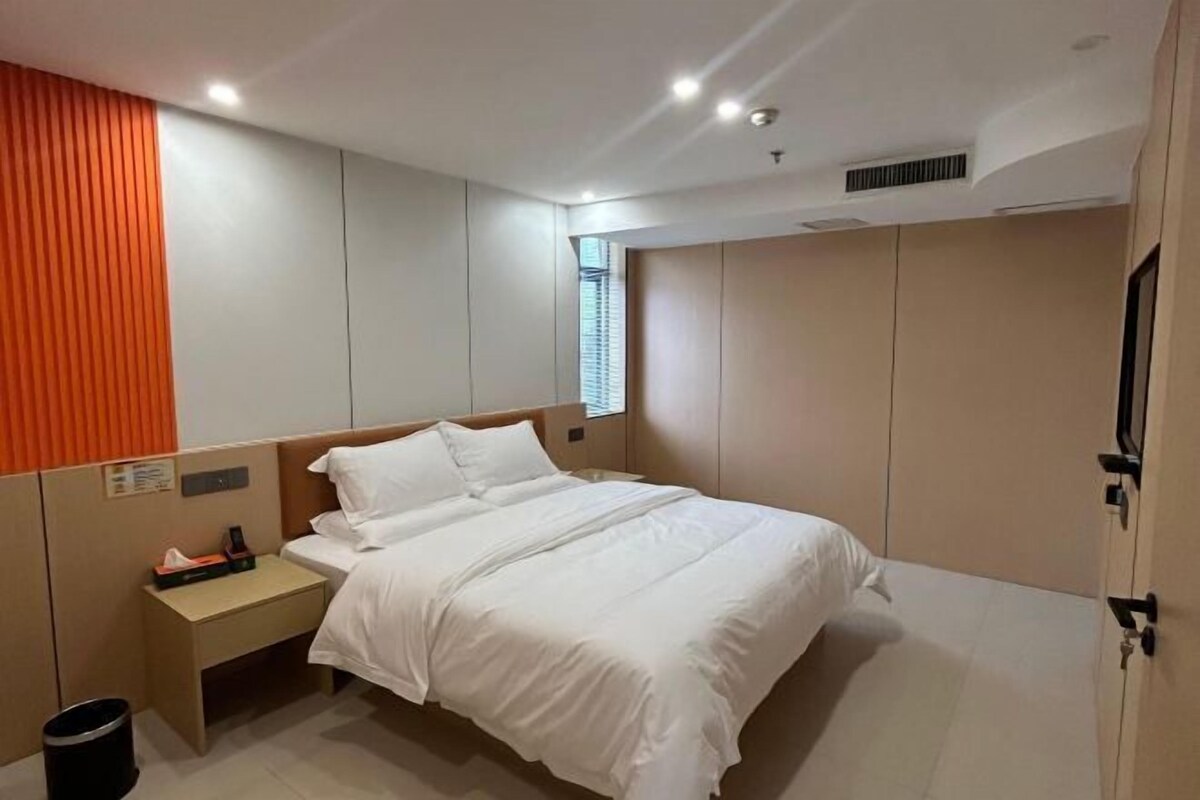 A modern hotel room with a large bed, a bedside table, a trash bin, and a window with blinds.