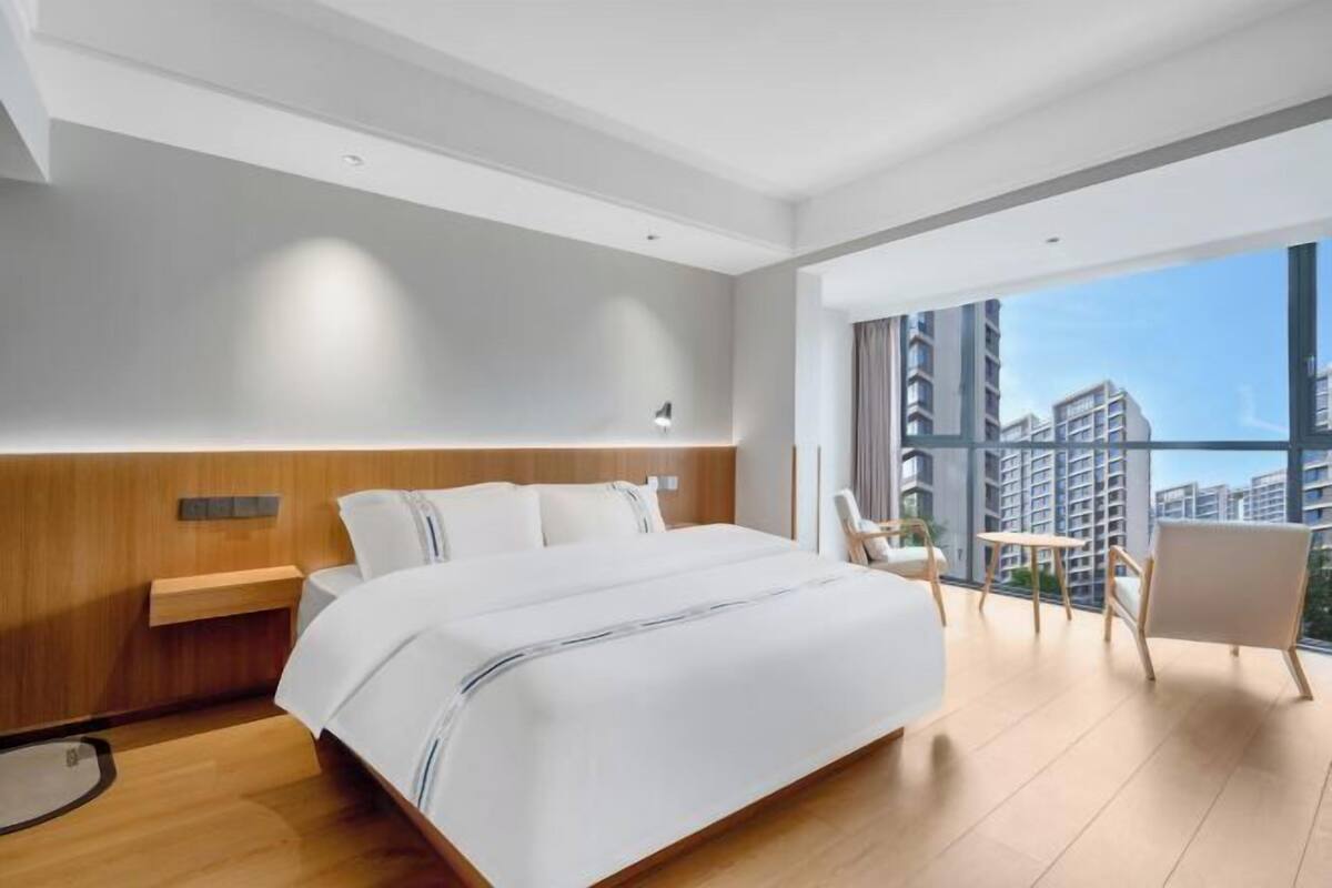 Quarto de hotel moderno com uma cama grande, uma mesa pequena e vista da cidade através de janelas amplas.