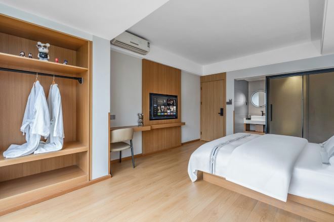 A modern hotel room with a large bed, a wooden wardrobe, a desk, and a bathroom with a mirror.