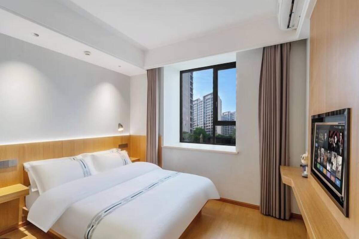 Quarto de hotel com uma cama grande, janela com vista para a cidade, televisão montada e cabeceira de madeira.