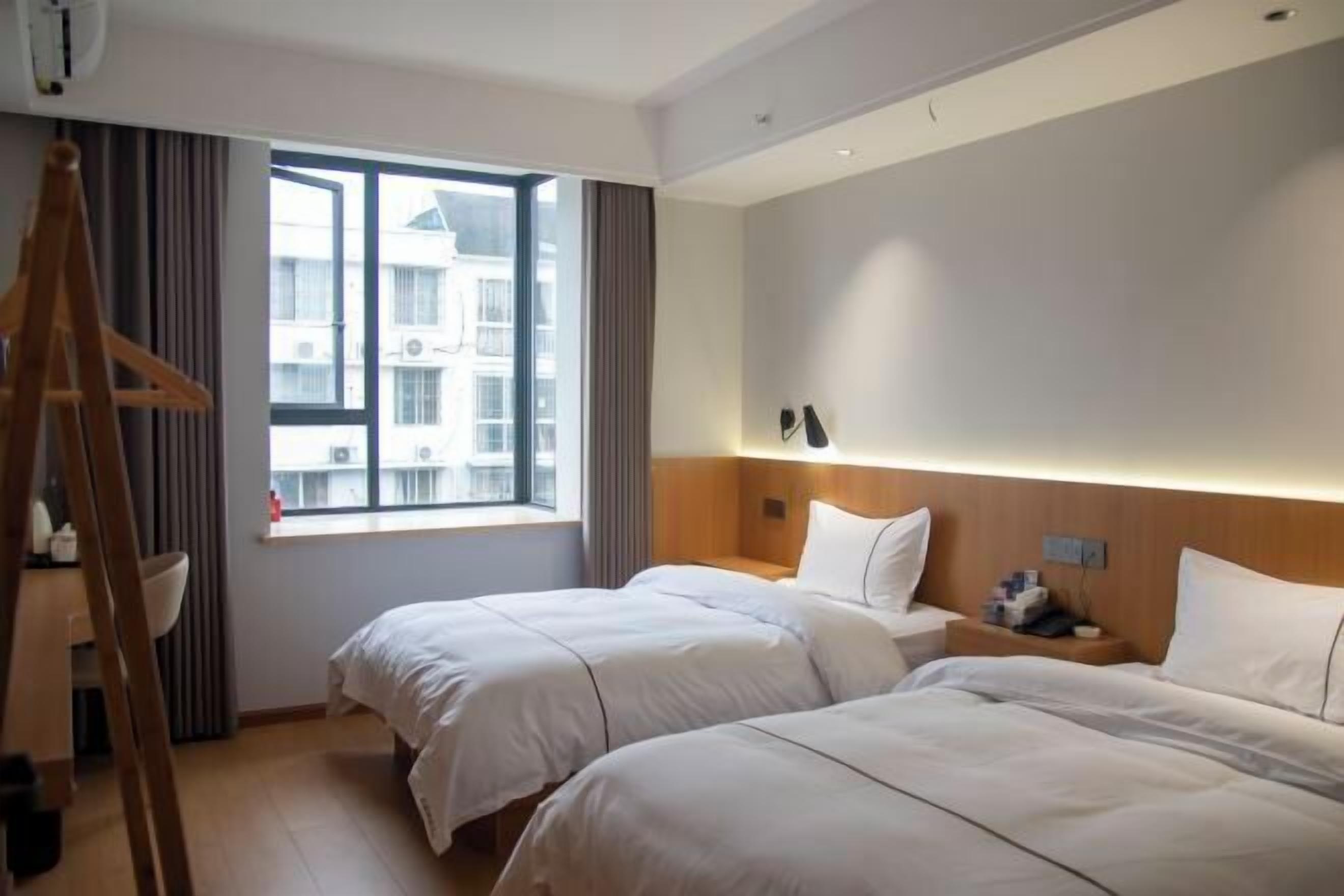 A hotel room with two beds, a wooden ladder, a window with a view of buildings, and a bedside table with a lamp.