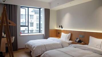 A hotel room with two beds, a wooden ladder, a window with a view of buildings, and a bedside table with a lamp.