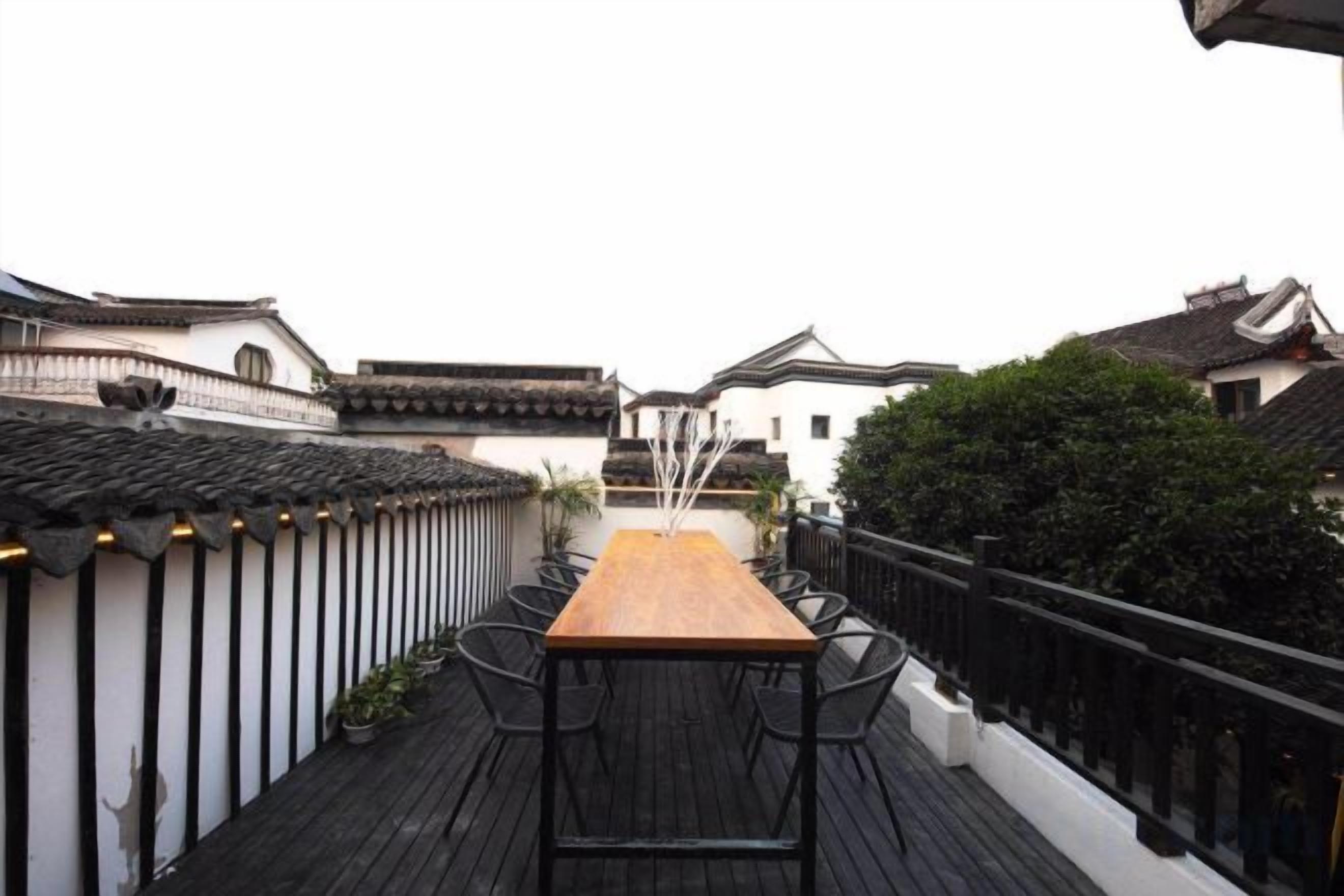 Una terraza de madera tradicional con una larga mesa y sillas, rodeada de edificios blancos con barandillas negras.