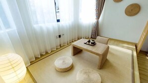 Ein Zimmer mit Tatami-Boden, einem niedrigen Tisch, zwei Kissen, einer Lampe und einem Fenster mit durchsichtigen Vorhängen.