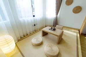 Une pièce avec un sol recouvert de tatamis, une table basse, deux coussins, une lampe et une fenêtre ornée de rideaux légers.