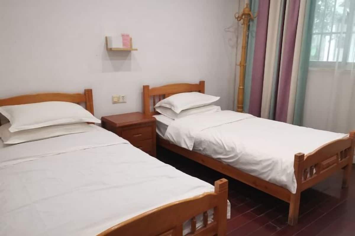 A room with two wooden beds, a bedside table, a shelf with a book, and a window with curtains.