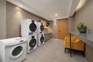 Zona de lavandería con lavadoras apiladas, una kitchenette pequeña y un área de descanso con una silla.