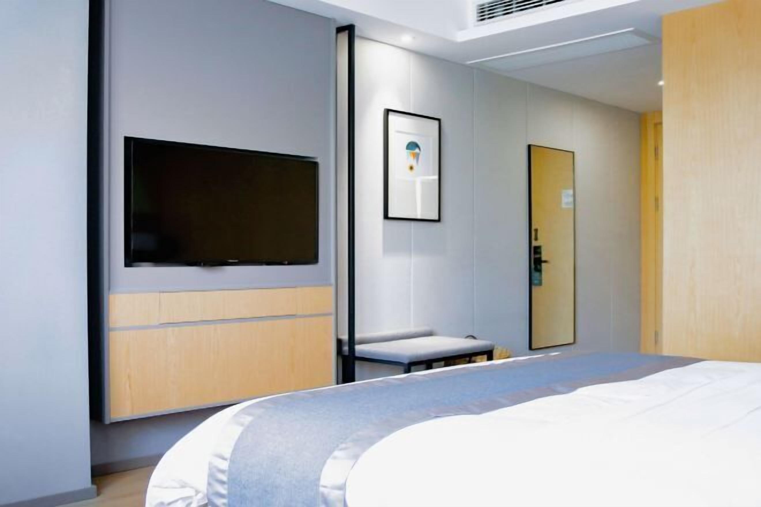 A modern hotel room with a bed, a mounted TV, a framed picture on the wall, and a door with a mirror.
