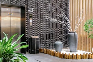 An elevator with a metallic finish, a wall with a geometric pattern, and a large grey vase with branches. - Free Living Hotel (Daishan)