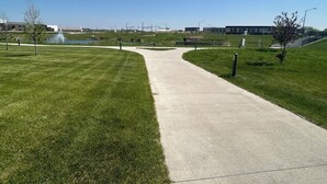 A paved pathway in a park with grass on either side, a fountain, and modern buildings in the background.