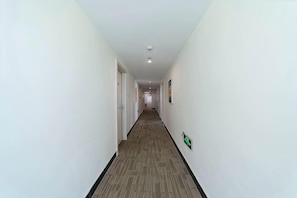 A long, narrow hallway with white walls and a carpeted floor, leading to a door at the end.