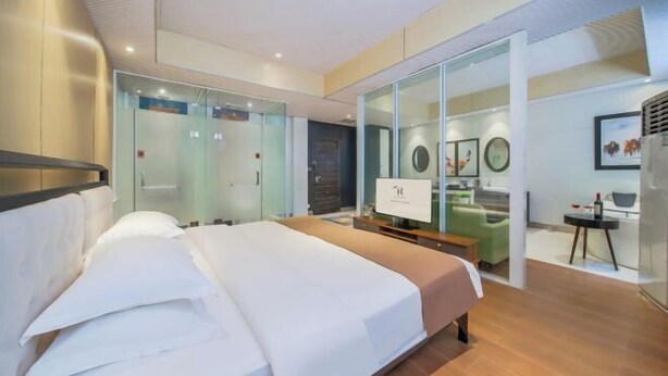 Une chambre d’hôtel moderne dotée d’un grand lit, d’un bureau et d’une cloison en verre qui sépare la chambre à coucher du coin salon.