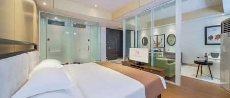 A modern hotel room with a large bed, a desk, and a glass partition separating the room from the hallway.