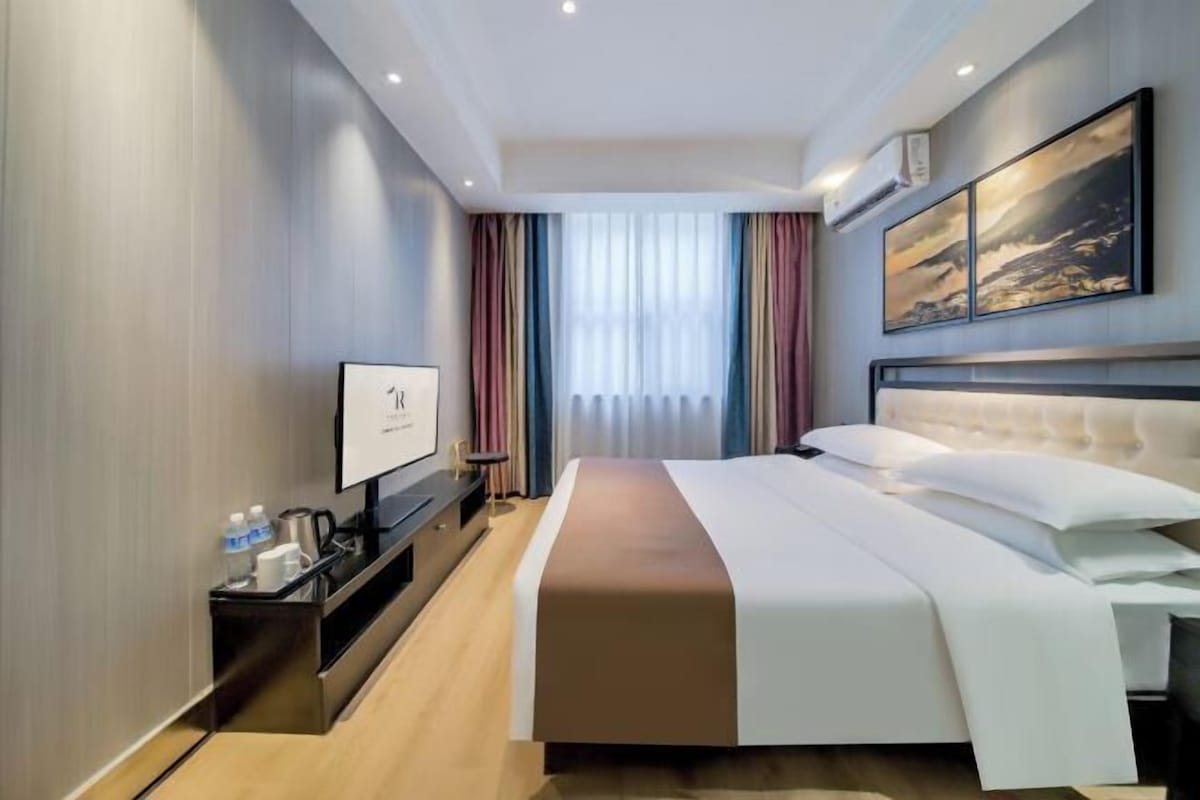 Ein modernes Hotelzimmer mit einem großen Bett, einem Flachbildfernseher, einem Nachttisch mit Kaffeemaschine und einem fenster mit Vorhängen.
