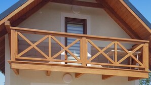 Un balcon en bois doté d’une simple balustrade, adossé à un bâtiment avec un toit en pente et une petite fenêtre.