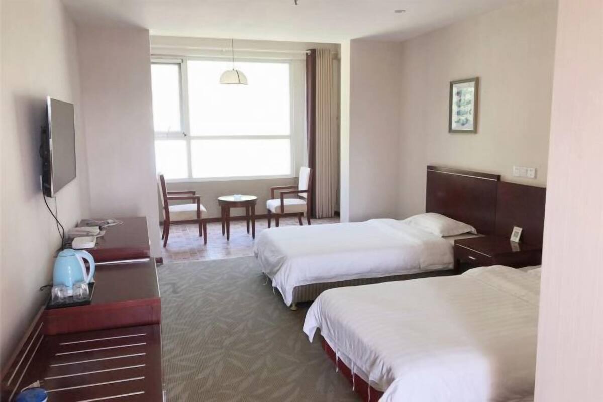 Ein Hotelzimmer mit zwei Betten, einem Schreibtisch mit Wasserkocher, einem Sessel, einem kleinen Tisch und einem Fernseher.