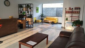 A modern lobby with a reception desk, a yellow sofa, a brown leather couch, a wooden coffee table, a plant shelf, and a large window with a view of a stone wall. - AA Hotel Chain Lianyungang Jinxu Garden Branch (Lianyungang)