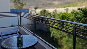 Un balcón con mesa y sillas, vistas a la vegetación y colinas al fondo, y un cielo despejado.