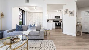 A modern living room with a grey sofa, a coffee table, and a patterned rug. A kitchen is visible in the background with white cabinets and stainless steel appliances.