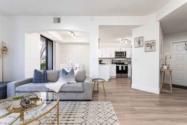 A modern living room with a grey sofa, a coffee table, and a patterned rug. A kitchen is visible in the background with white cabinets and stainless steel appliances.