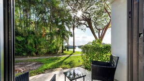 An outdoor patio with two black wicker chairs and a small table, surrounded by lush greenery and a view of a body of water.