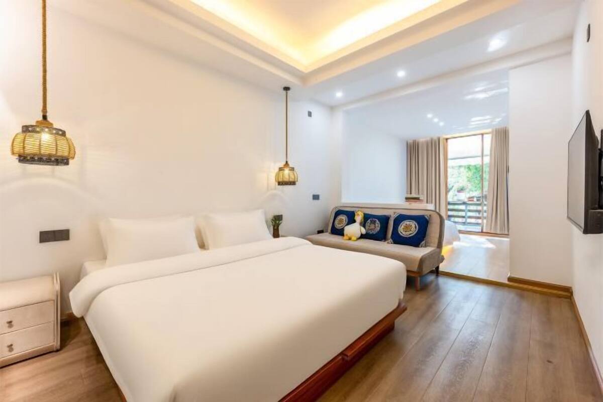 A bedroom with a large bed, bedside tables, a sofa with cushions, a TV, and a window with curtains.