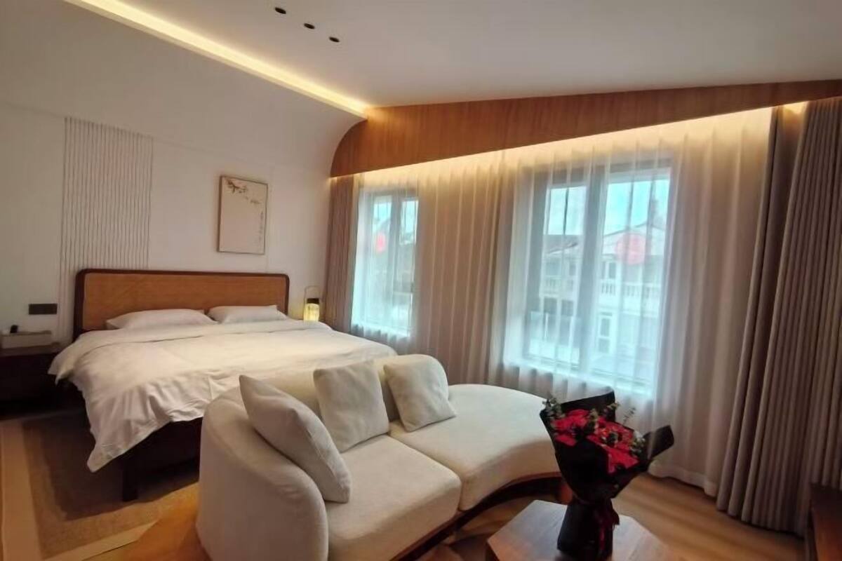 A hotel room with a bed, a sofa, a bedside table, a lamp, and a window with curtains.