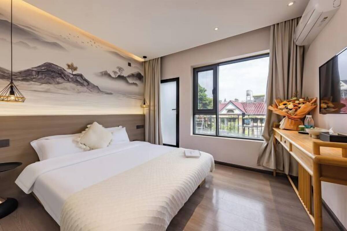 A hotel room with a bed, a desk, a window with a view, and a mural on the wall.
