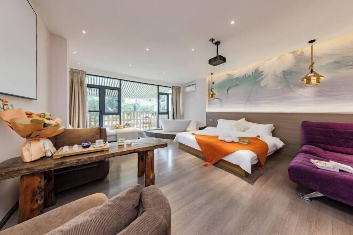 A modern hotel room with a bed, sofa, dining table, and a large window.