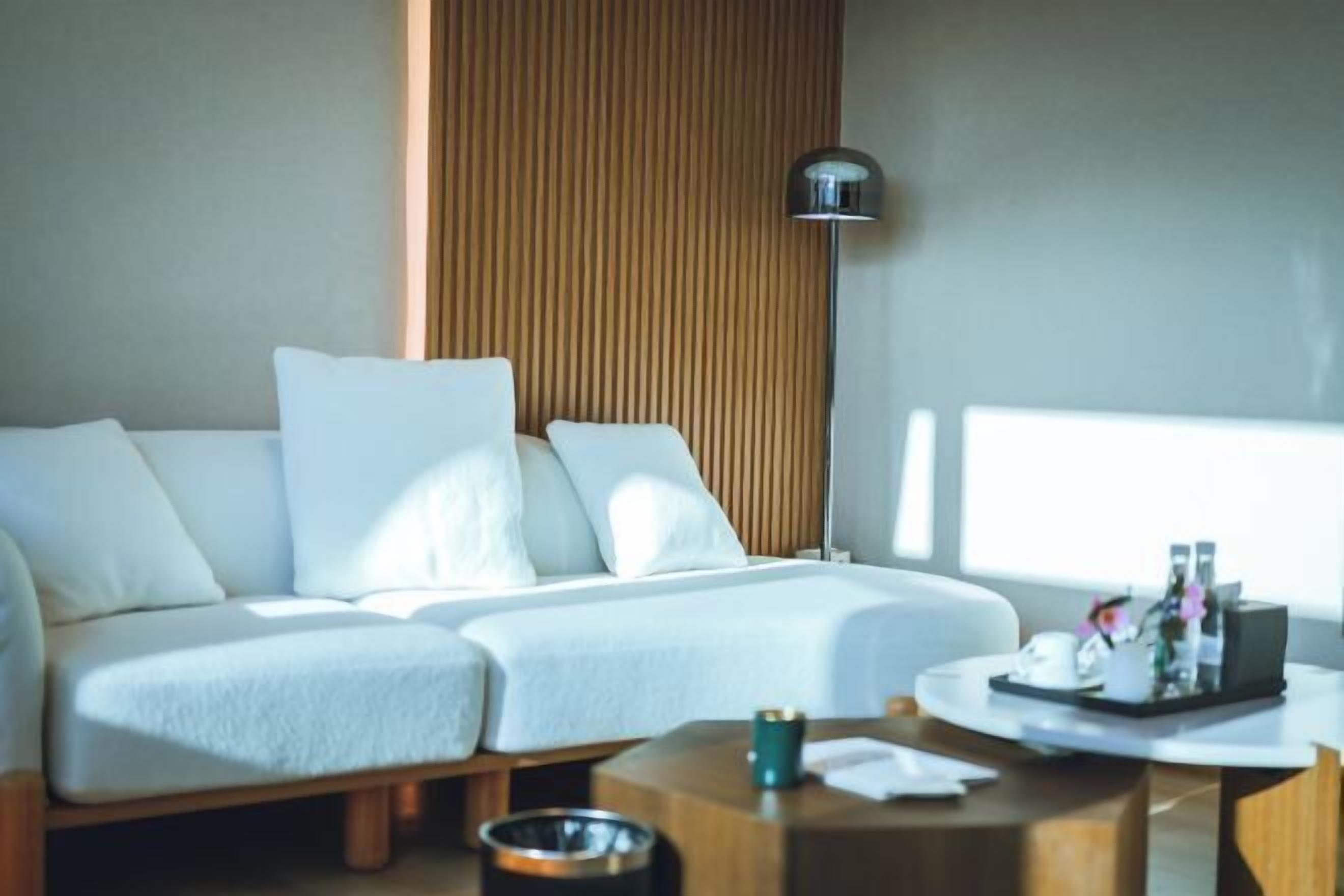 A modern hotel room with a white sofa, a wooden accent wall, a floor lamp, and a small table with a tray of toiletries.