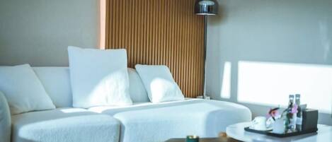 A modern hotel room with a white sofa, a wooden accent wall, a floor lamp, and a small table with a tray of toiletries.