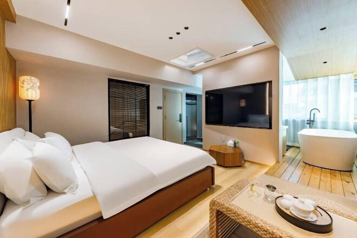 A modern hotel room with a large bed, a flat-screen TV, a bathtub, and a wooden floor.