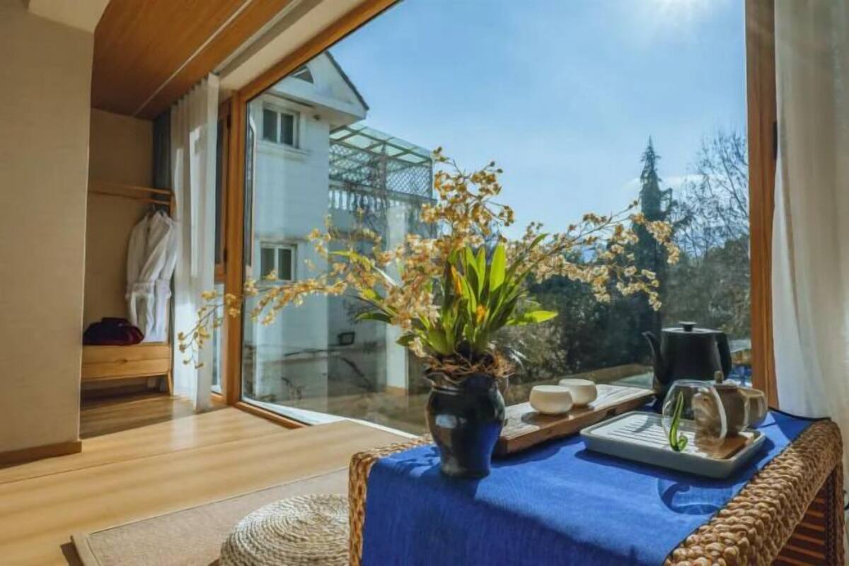 A room with a large window, a wicker sofa, a table with a blue tablecloth, a potted plant, and a kettle.