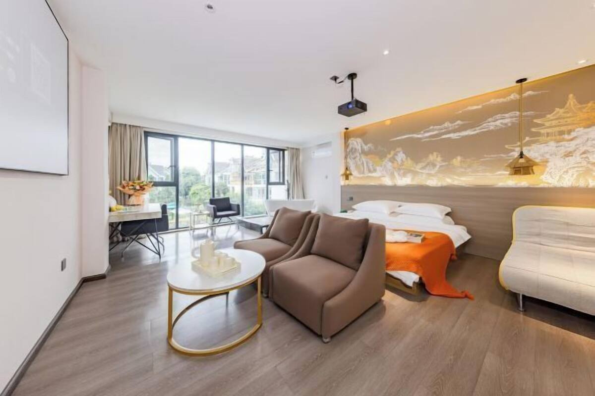A modern hotel room with a large window, a sofa, a bed with an orange accent, and a dining area with a table and chairs.