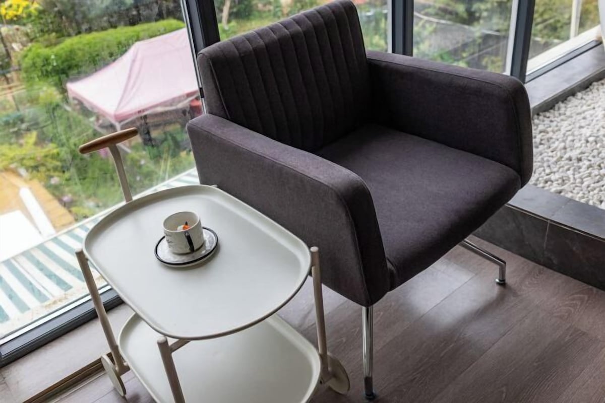 A modern room with a dark upholstered chair, a small white table with a cup, and a view of greenery outside.