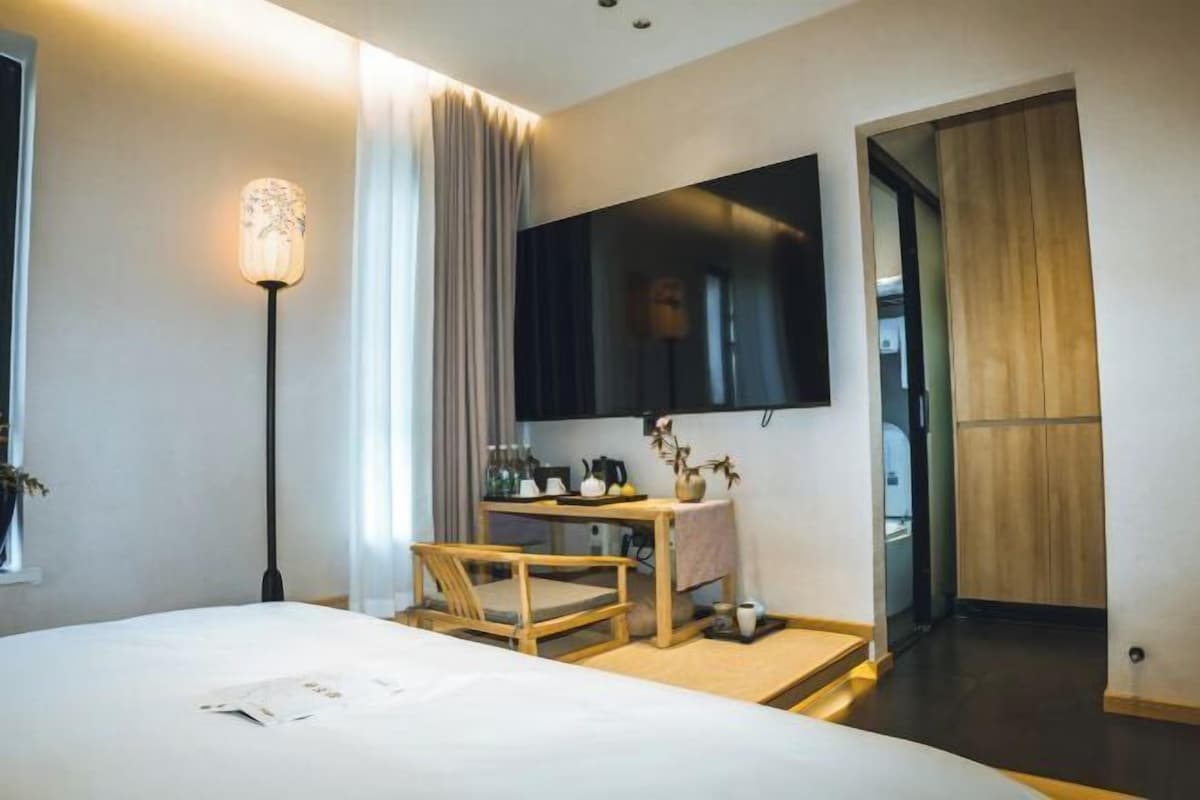 A modern hotel room with a large bed, a desk with a chair, a flat-screen TV mounted on the wall, and a wooden wardrobe.
