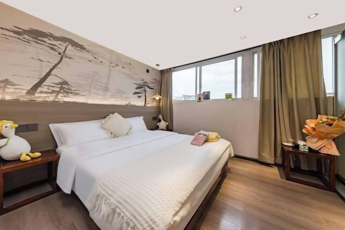 A hotel room with a bed, a nightstand, a wall mural, a window with curtains, and a small table with a bouquet.