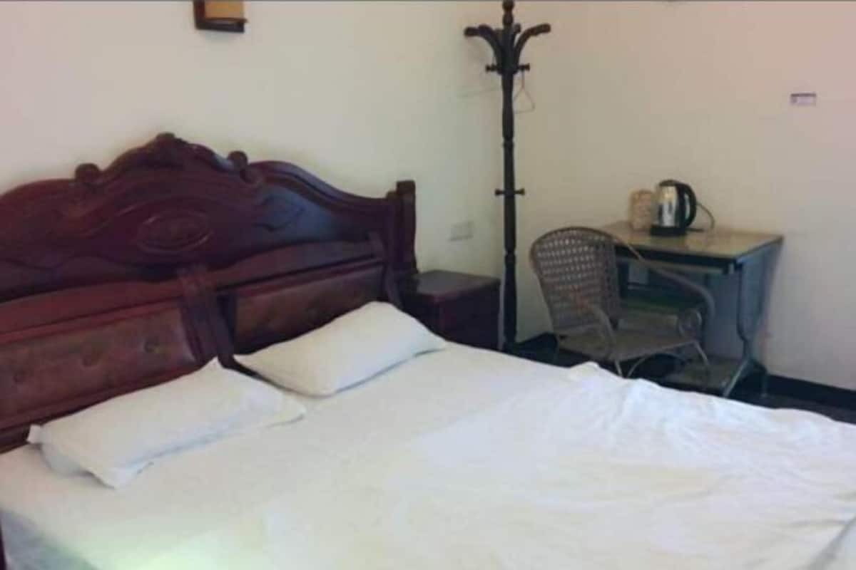 Ein Bett mit weißer Bettwäsche, ein Holz-Kopfstück, ein kleiner Schreibtisch mit Stuhl und eine Stehlampe.