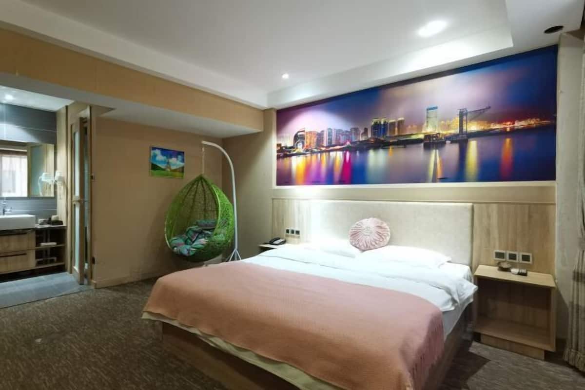 A hotel room with a bed, a hanging chair, a nightstand, and a large cityscape painting on the wall.