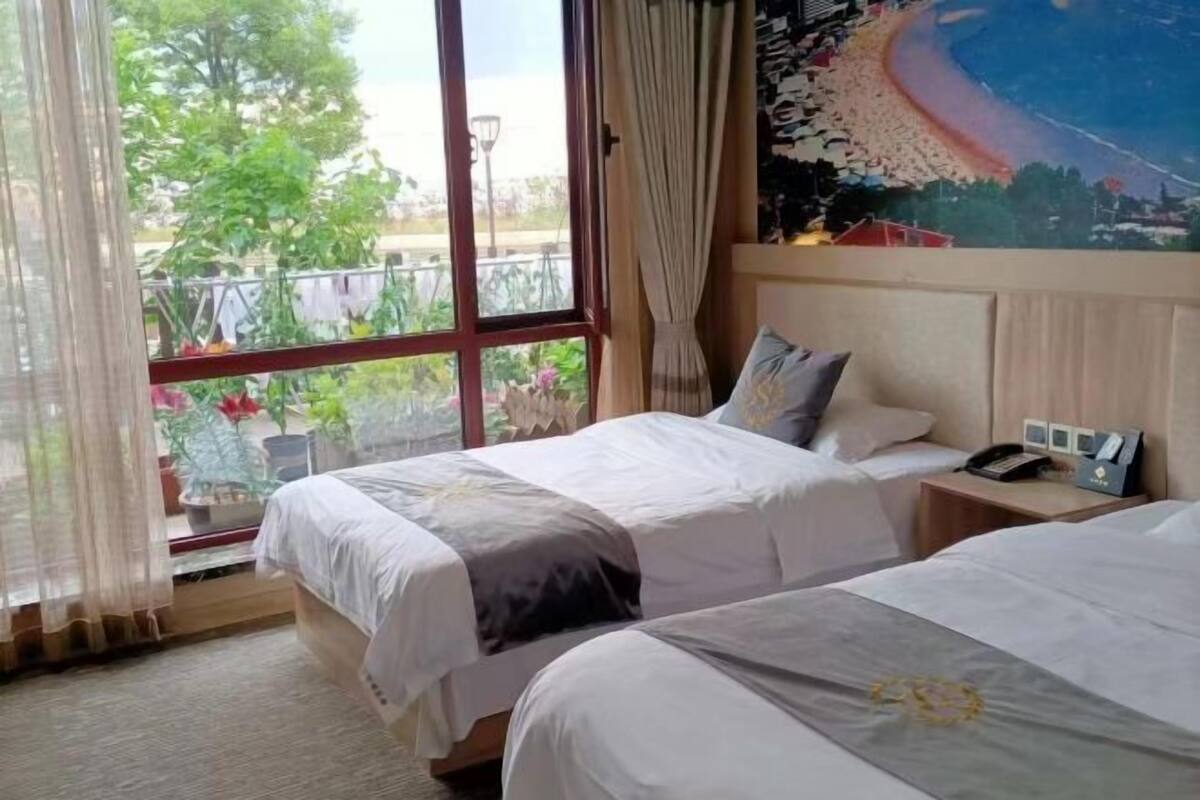 A hotel room with two beds, a large window with curtains, a painting on the wall, and a small table with a phone and a box.