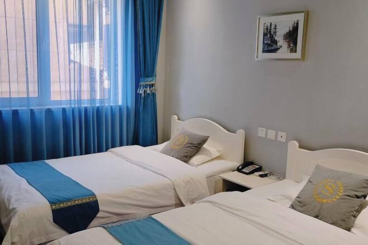 A hotel room with two single beds, a nightstand with a phone, and a window with blue curtains.