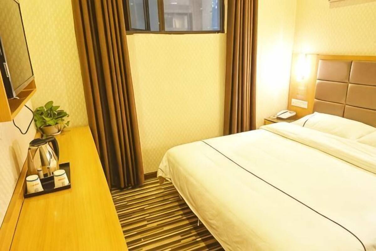A hotel room with a bed, a bedside table with a plant and a tray of items, a window with curtains, and a wall-mounted TV.