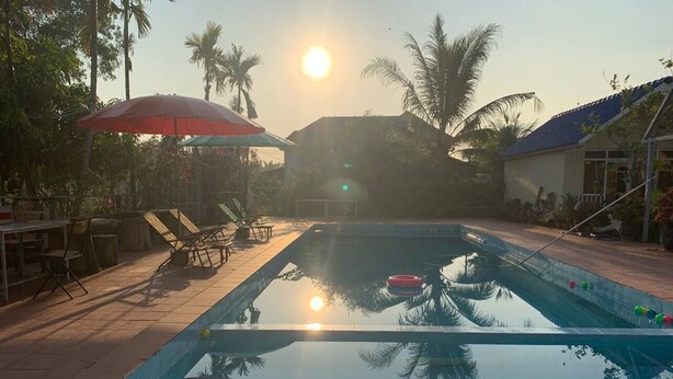 A swimming pool with a clear view of the sun setting in the background, surrounded by palm trees and outdoor seating. - Ratana Bungalow (Kampot)