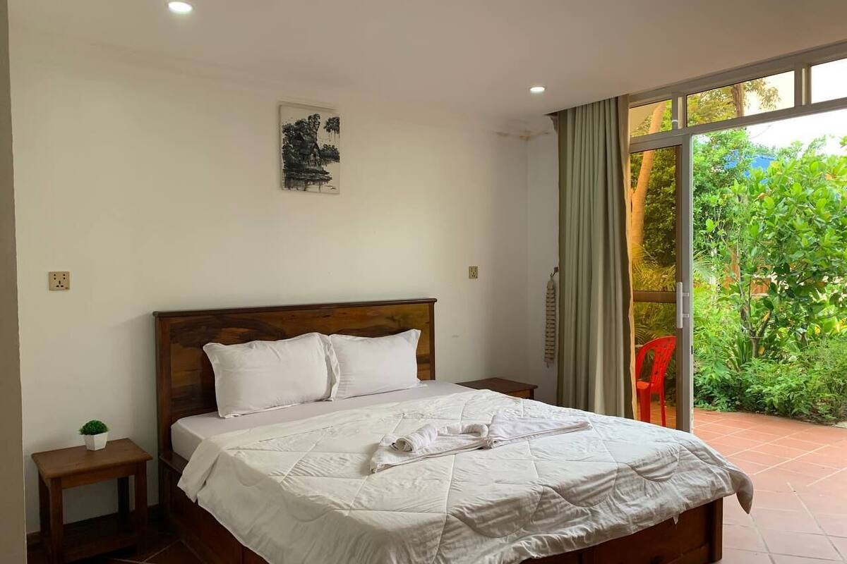 Una cama bien hecha con sábanas blancas y una toalla doblada, una mesita de noche con una planta, y una ventana con cortinas.